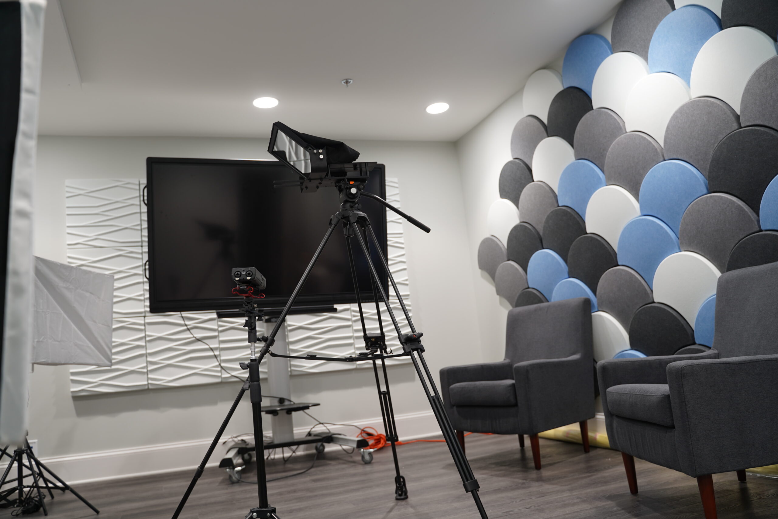A video production setup in a modern room with a camera on a tripod facing a flat-screen TV, a softbox light to the left, two armchairs to the right, and a decorative wall with colorful panels.