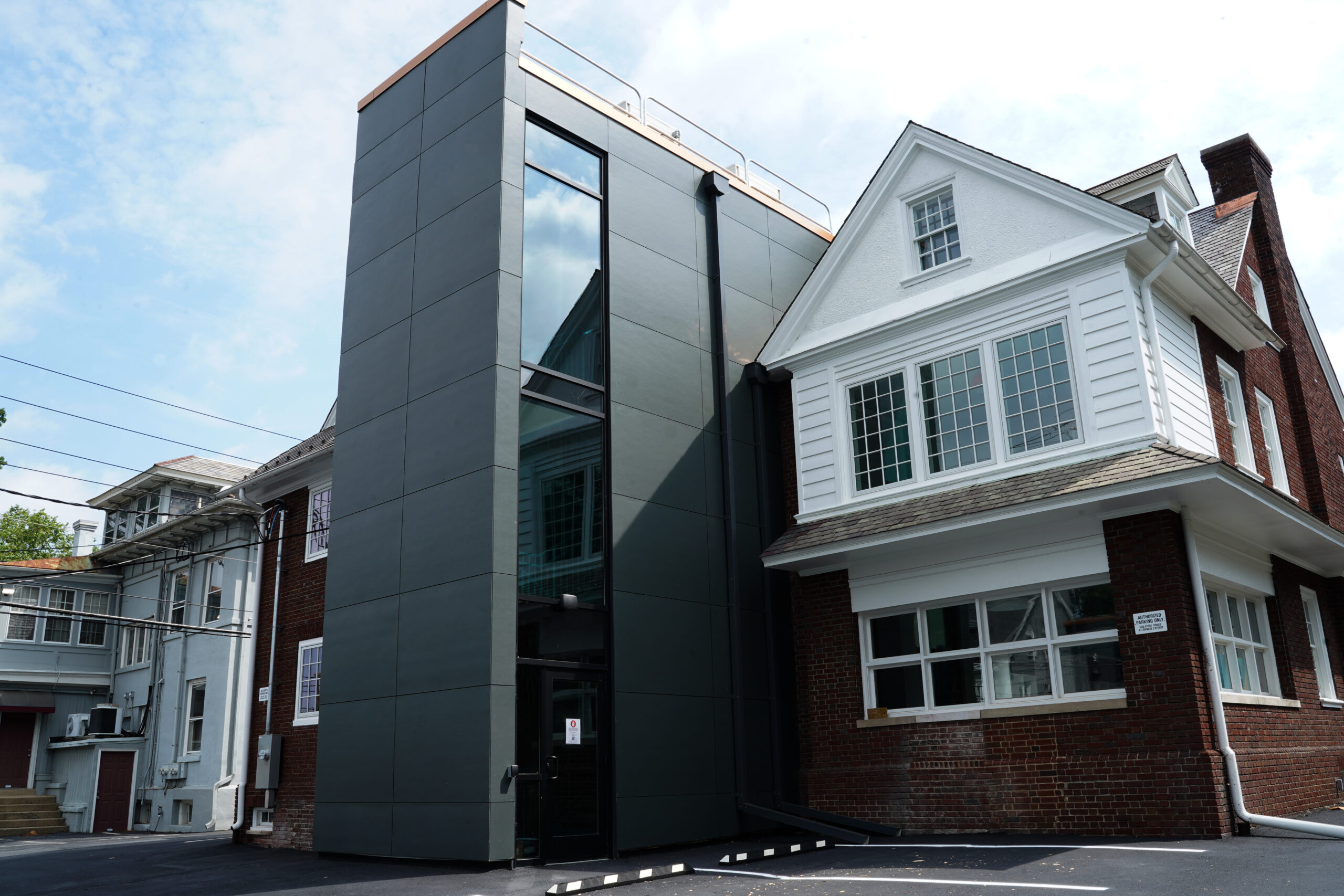 A modern dark-colored building extension with large windows attached to a traditional white house with a gabled roof and brick foundation under a partly cloudy sky.