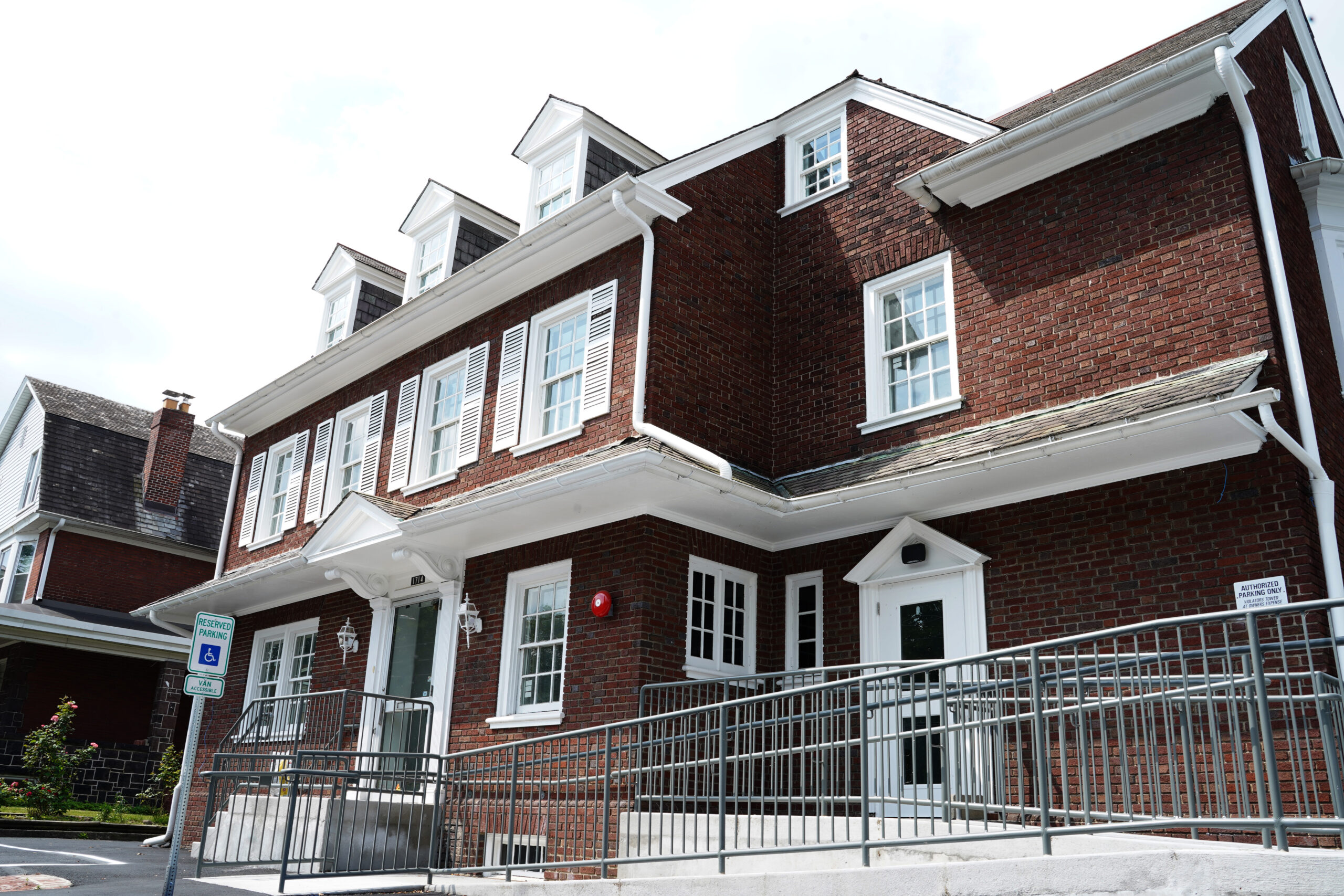 A two-story brick building with white trim and dormer windows, featuring a small porch with a metal railing and signs for reserved parking and authorized personnel only.