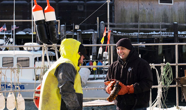 Two men at a snowy harbor, one in a yellow fisherman's outfit and the other holding a lobster, with boats in the background.