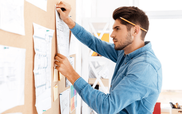 Focused young man with a pencil behind his ear attaching a document to a corkboard covered with various notes and papers.