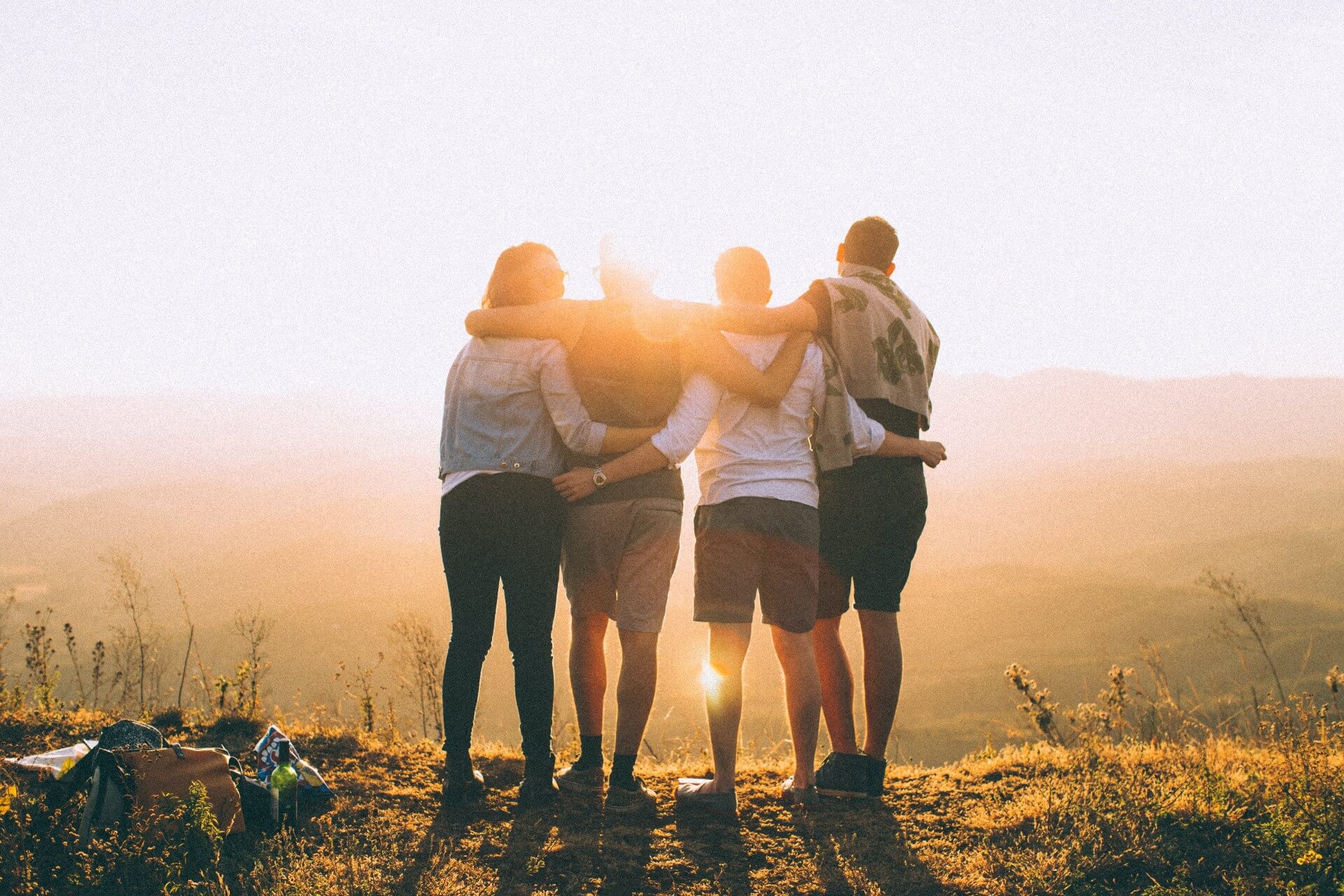 Four friends standing together with arms around each other, looking at a sunset over a hilly landscape, with personal items on the ground, suggesting an outdoor adventure.