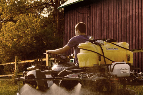 Person operating a riding lawn mower with a CropCare tank, spraying a substance on the ground near a red barn in a rural setting during dawn or dusk.