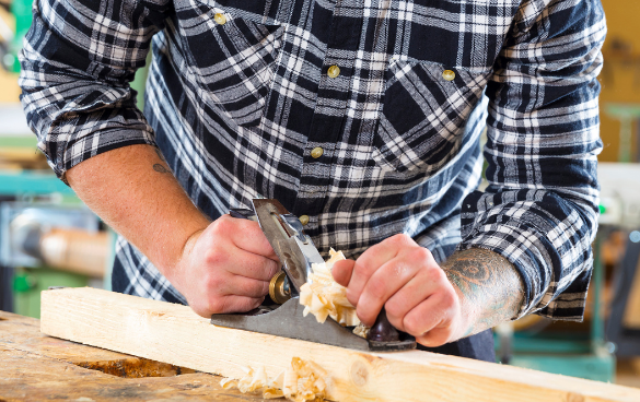 A person in a plaid shirt using a hand plane on a piece of wood in a workshop, with visible tattoos on their arms.