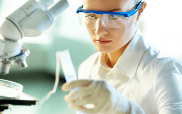 A female scientist in safety goggles and a lab coat examines a slide in a laboratory with a microscope in the background.