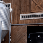 A wooden barn with a partially open sliding door next to a metallic grain silo, with the front of a vintage truck peeking out from the barn's interior.