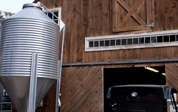 A wooden barn with a partially open sliding door next to a metallic grain silo, with the front of a vintage truck peeking out from the barn's interior.