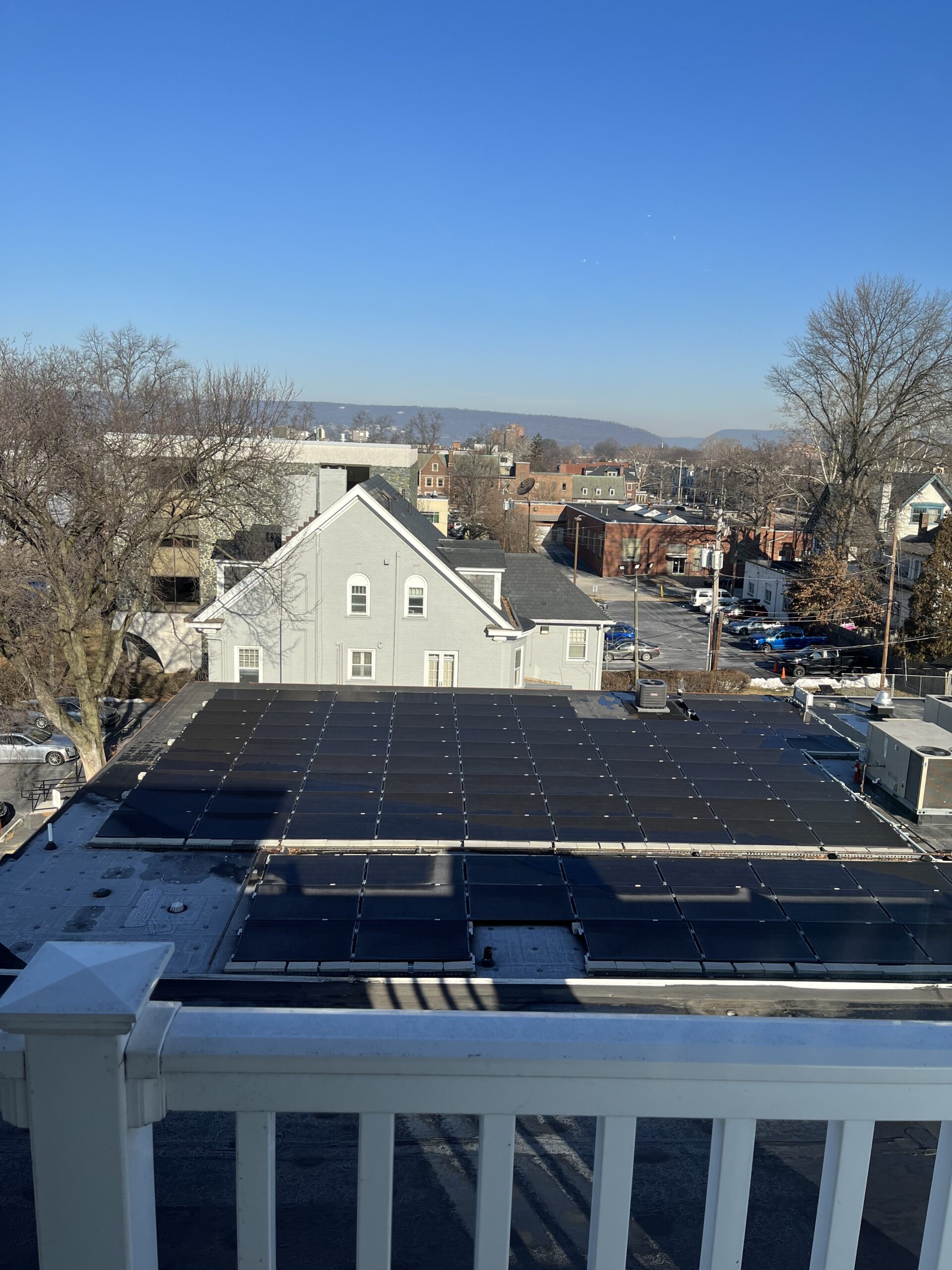 View from a balcony with a white railing overlooking a roof with solar panels, with houses, buildings, and distant hills under a clear blue sky.