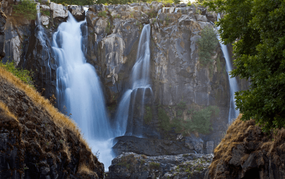 A powerful waterfall flowing over a rocky cliff surrounded by green vegetation, with a smooth, silky appearance to the water due to a long exposure photograph.