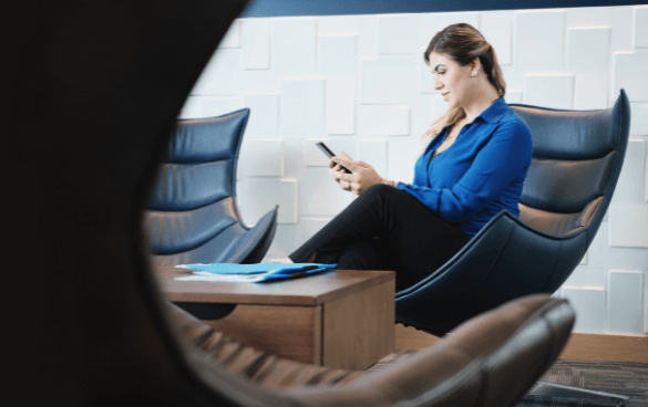 A woman in a blue blouse sitting in a modern chair, looking at her smartphone with a geometric patterned wall in the background.