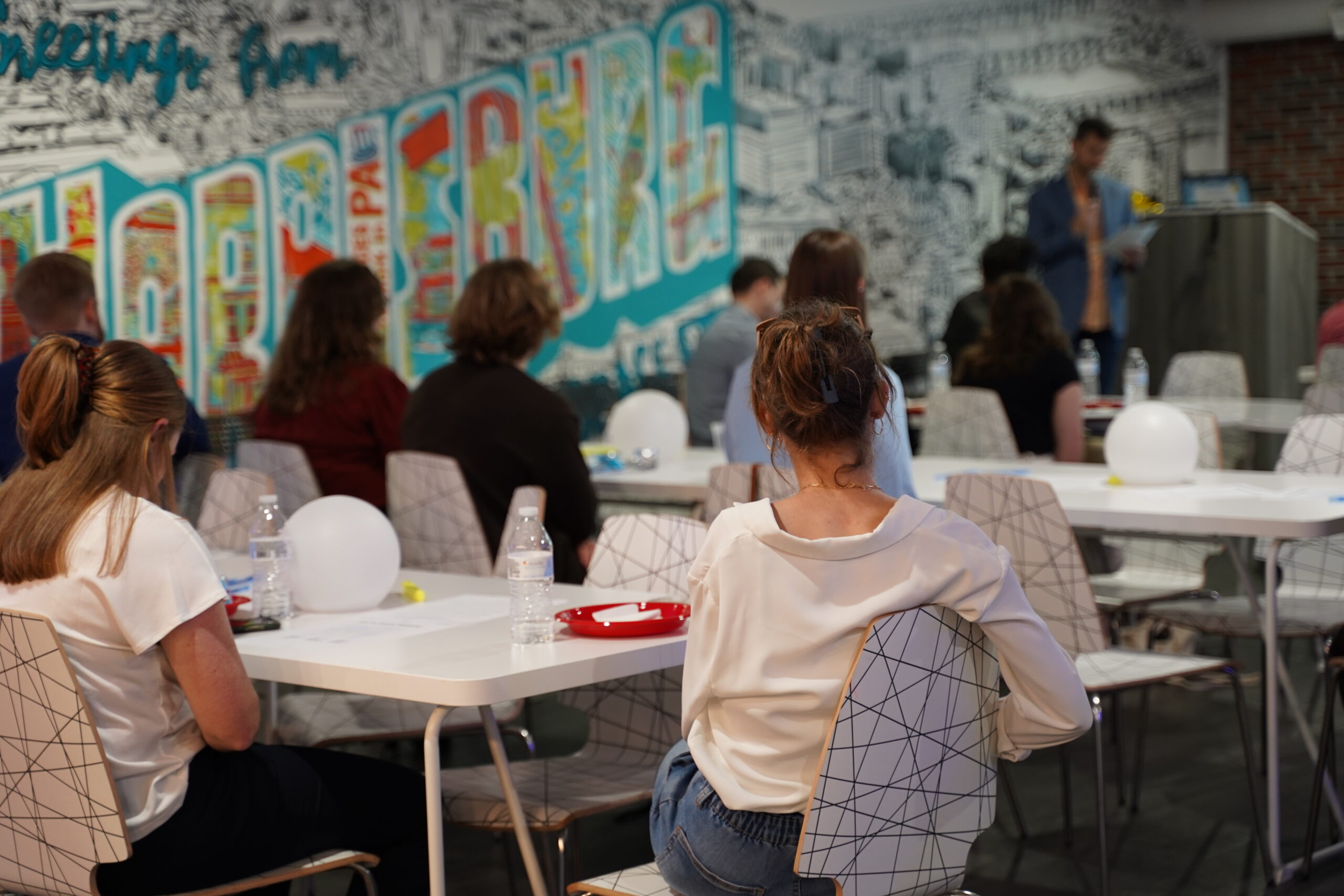 Attendees seated at tables listening to an out-of-focus presenter in a room with a colorful mural, during a workshop or seminar.