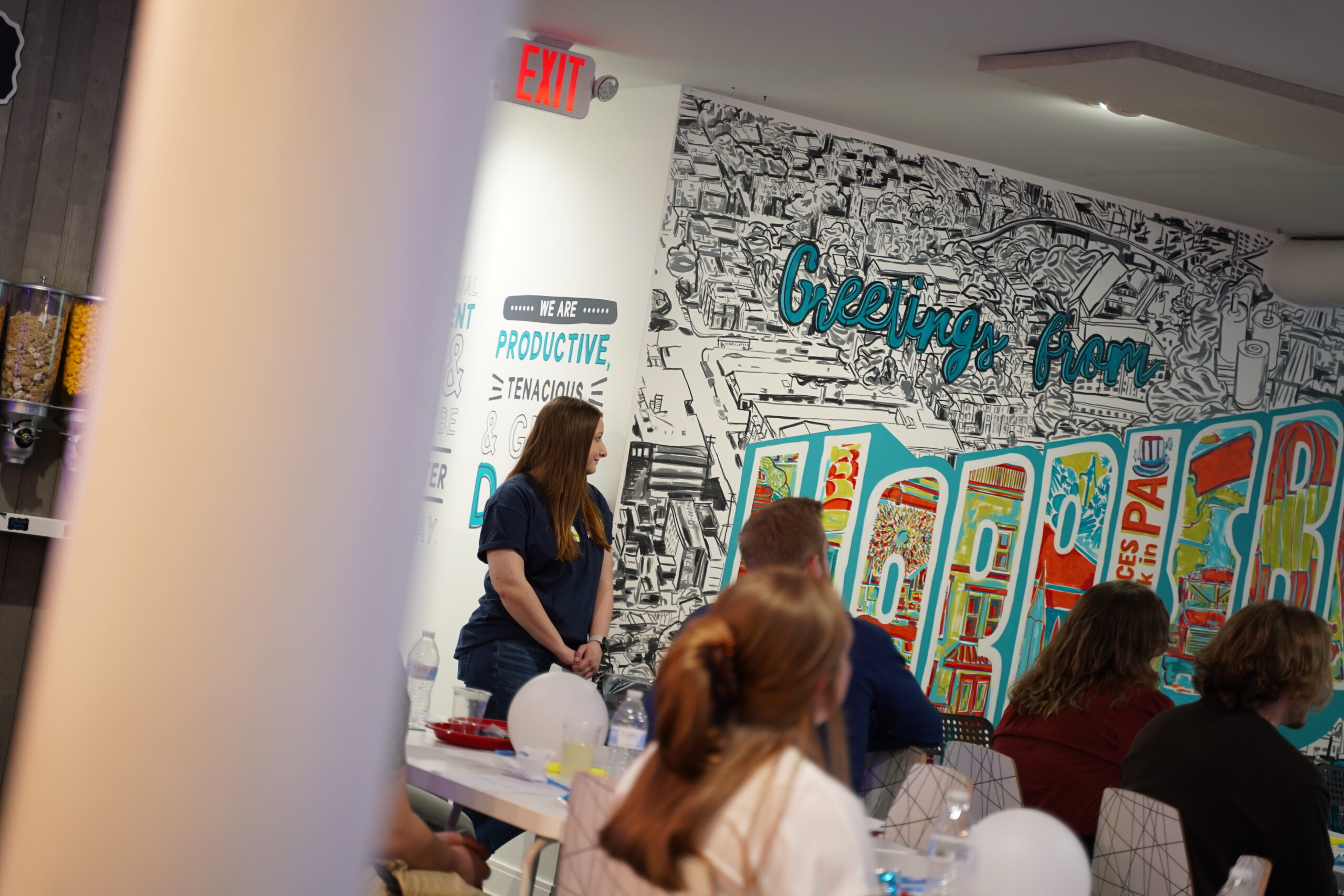 People gathered in a room with a colorful ice cream-themed mural on the wall, engaging in conversation at tables, with a popcorn machine on the left.