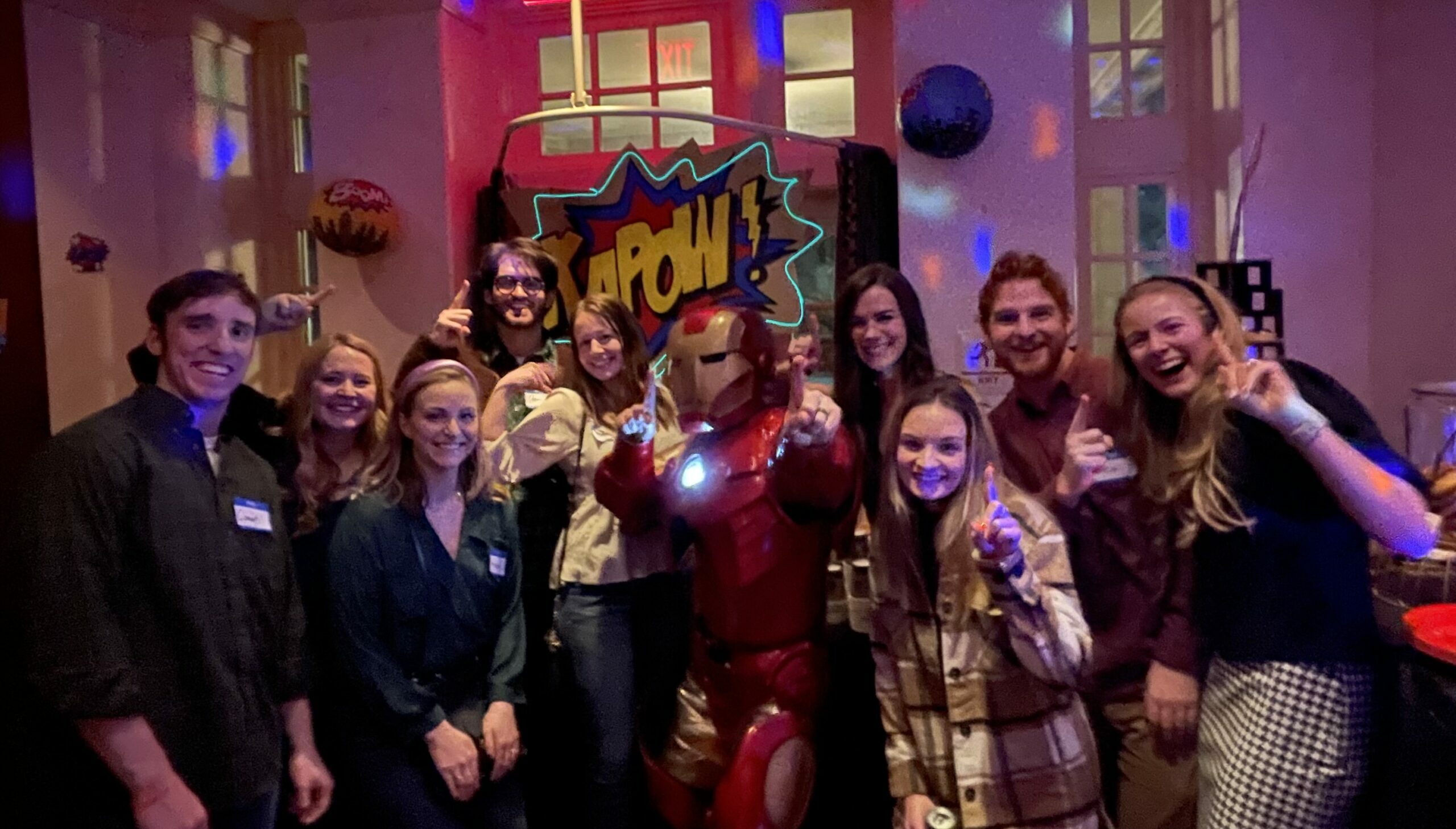 A group of nine people posing with a person in an Iron Man costume at a superhero-themed party, with comic book-style decorations in the background.