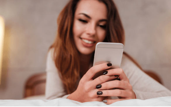 A smiling woman lying down while looking at her smartphone, with black nail polish on her nails, in a cozy indoor setting.