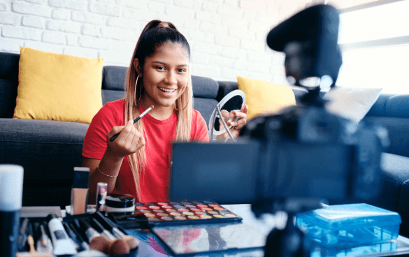 A young woman records a makeup tutorial, applying makeup while looking into a handheld mirror, with a camera on a tripod capturing the session and makeup products spread out on a table in front of her.