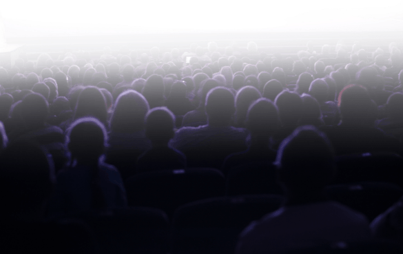 Blurred silhouettes of an audience in a dark theater facing a bright light where a screen would be.