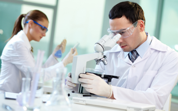 Two scientists in a lab, with a man using a microscope and a woman examining a test tube, both wearing safety goggles and white lab coats.