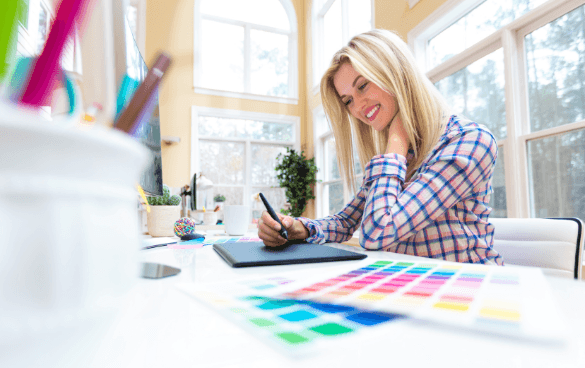 A smiling woman looking at her smartphone while sitting at a desk with art supplies and a color swatch palette in a sunny room.