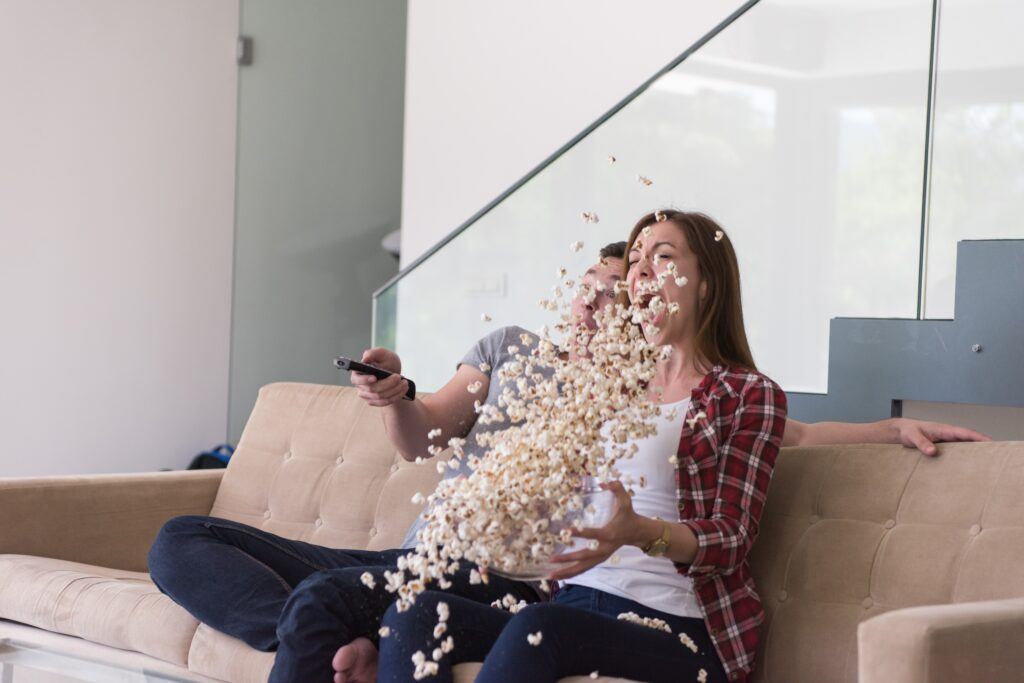 Two people on a sofa with popcorn flying in the air; the man holds a remote control and the woman laughs as she spills the popcorn.