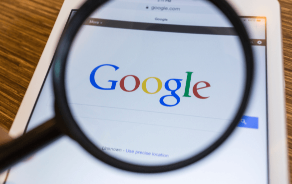 A magnifying glass held over a tablet screen displaying the Google homepage, focusing on the Google logo.