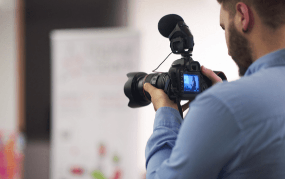 A person from behind operating a professional video camera with a microphone, focusing on a subject out of view, with a blurred whiteboard in the background.