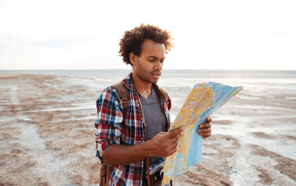 A young man with curly hair, wearing a plaid shirt and a camera around his neck, is reading a map outdoors on a vast, flat terrain with a clear sky.