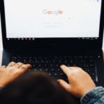 Person using a Dell laptop with Google search homepage on the screen, a smartphone, pen, and a potted plant with orange flowers on the table.