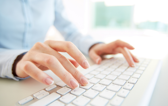 Close-up of a person's hands typing on a white keyboard, wearing a light blue shirt, with a blurred background.