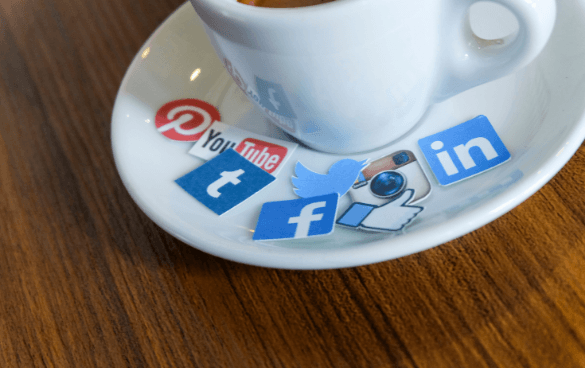 A white coffee cup on a saucer with paper cutouts of social media logos such as Pinterest, YouTube, Tumblr, Twitter, Facebook, and LinkedIn underneath it.