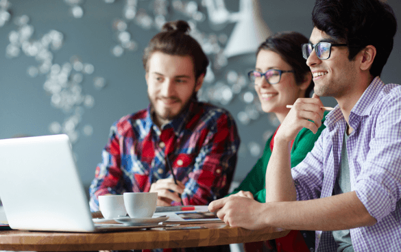 Three young adults collaborating around a laptop with coffee cups on the table in a creative workspace environment.