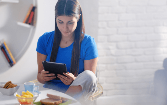 A woman in a blue shirt sitting at a table, focused on reading something on her tablet with snacks and a sandwich on the table.