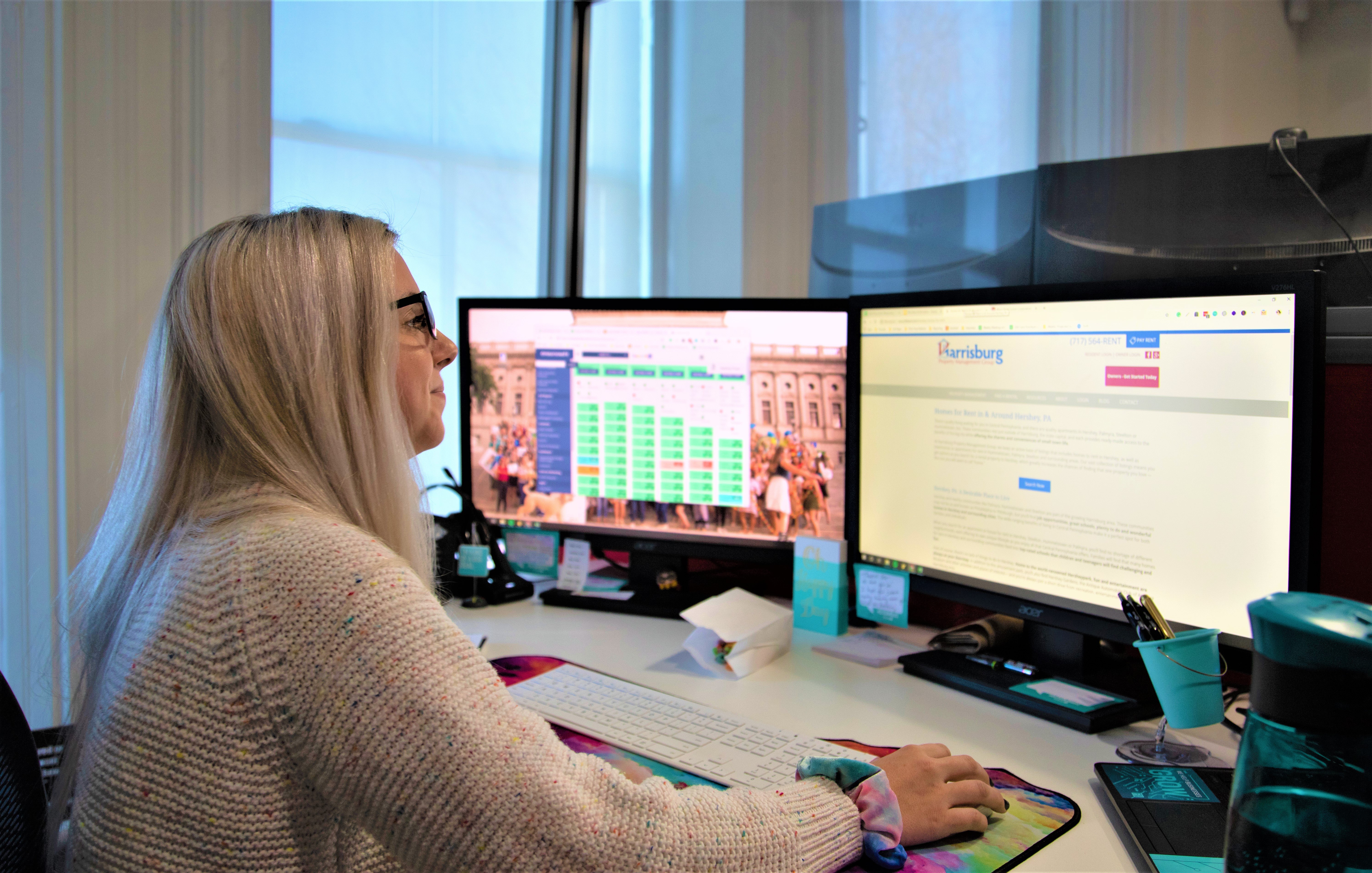 A woman with glasses working at a desk with two monitors displaying a spreadsheet and a Harrisburg webpage, surrounded by office supplies.