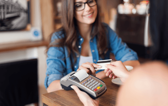 Smiling woman in a blue denim shirt accepting a credit card payment from a customer using a card terminal in an indoor setting.