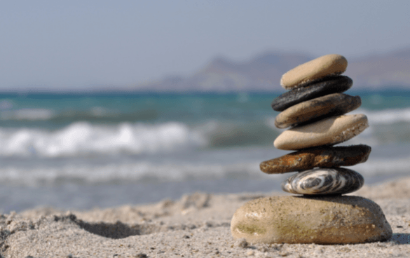 A stack of seven balanced stones on a sandy beach with a blurred background of a turquoise sea, waves, and distant mountains.