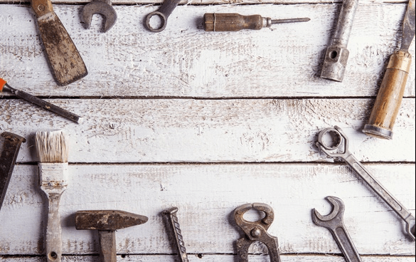 A collection of vintage tools including a hammer, wrenches, pliers, a screwdriver, a paintbrush, a chisel, and various nails and screws, displayed on a weathered white wooden surface.