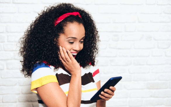 A smiling young woman with curly hair and a red headband looks at her smartphone with a hand on her cheek against a white brick wall background.