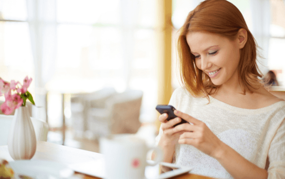 Smiling woman with reddish hair looking at her smartphone, sitting at a table with a white vase of pink flowers and a white mug in a bright room.