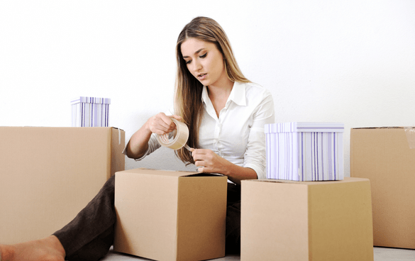 A woman sitting on the floor, taping up cardboard boxes, likely packing or unpacking items, in an indoor setting.