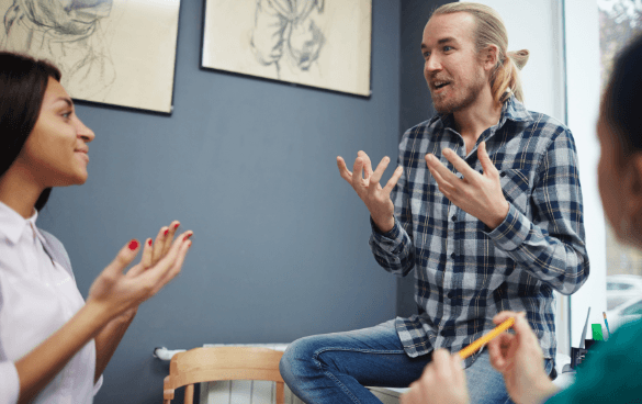 Three people engaged in a lively discussion, with a man in the center speaking and gesturing, a woman to his left responding with hand gestures, and a partial view of another person taking notes, in a room with artwork on the walls.