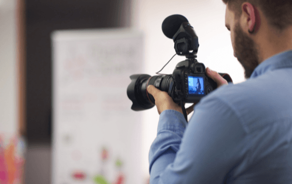 A camera operator from behind using a professional video camera to record a scene indoors, with a poster visible in the background.