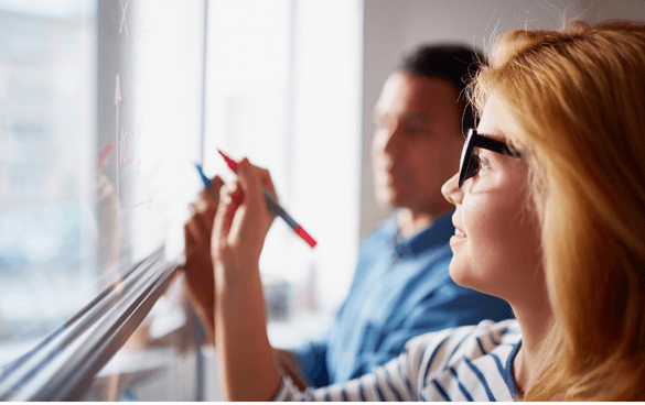 A woman with glasses writing on a clear board with a red marker as a man looks on, both engaged in a collaborative task.