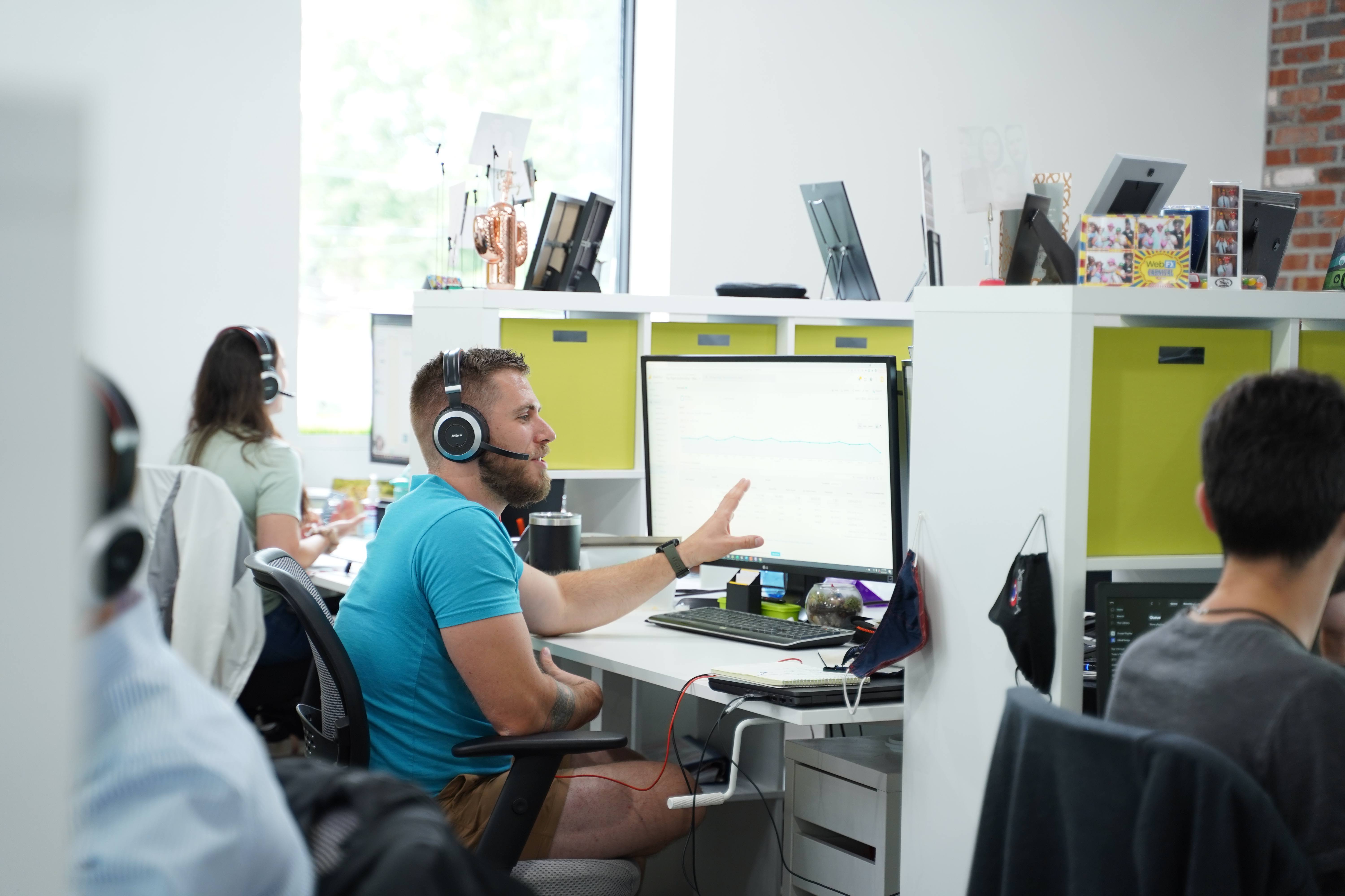 A man in a blue t-shirt and headphones gestures at a monitor with graphs in a modern office setting with other workers in the background.