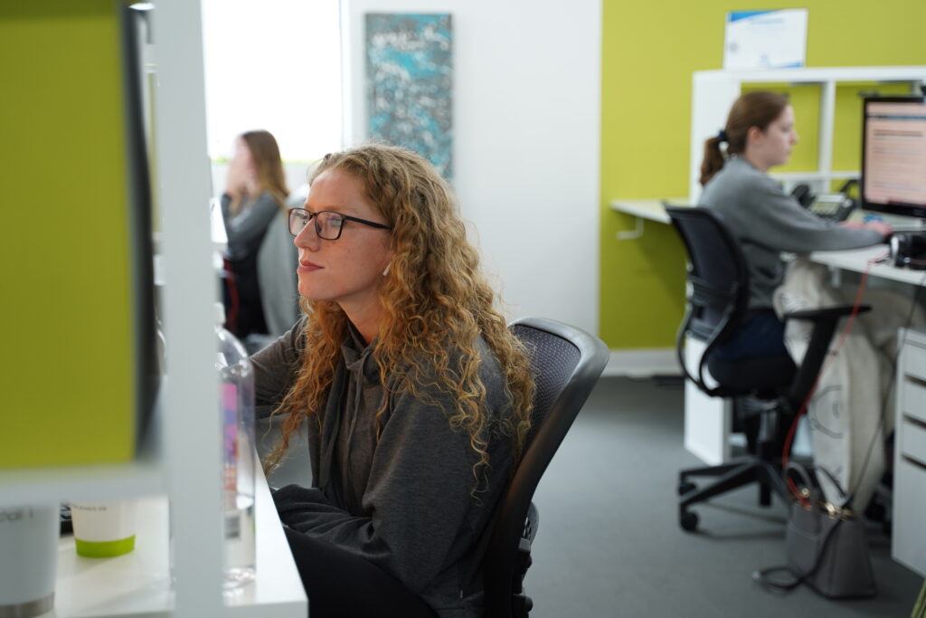 Two women working in a modern office setting, with the woman in the foreground looking at a computer screen and the woman in the background working on a dual monitor setup.