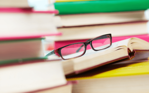 A pair of glasses resting on an open book with a stack of colorful books in the background.