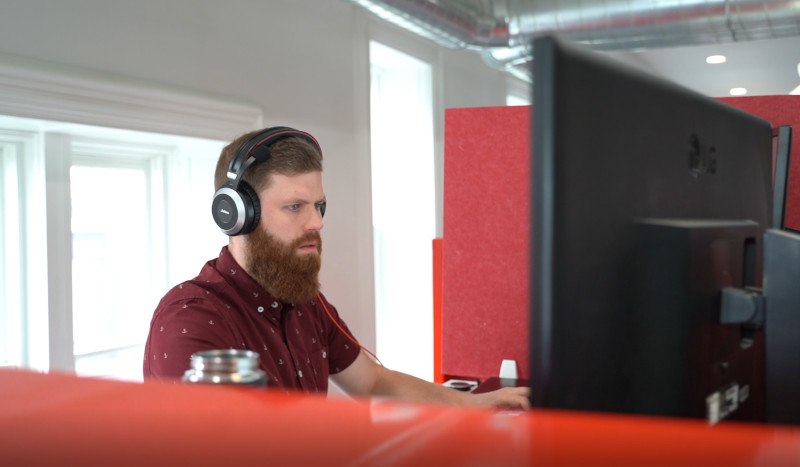 A man with a beard wearing headphones working intently at a computer in an office with red acoustic panels.