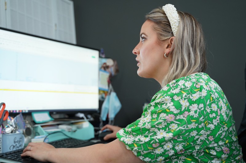 A woman with a green floral dress and a pearl headband is working at a desk with a computer and various office supplies.