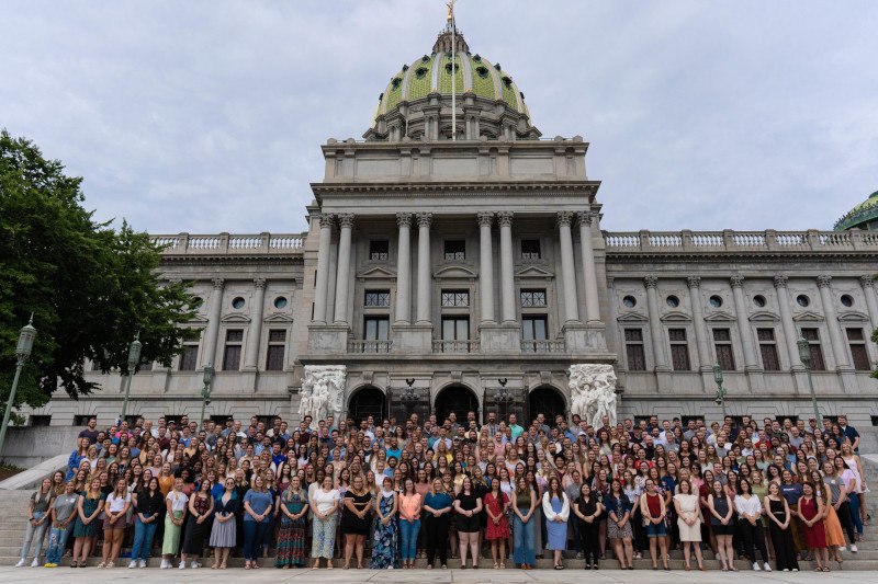 A large group of people posing for a photo on the steps of a grand building with a green-domed roof, likely a government or state capitol building.