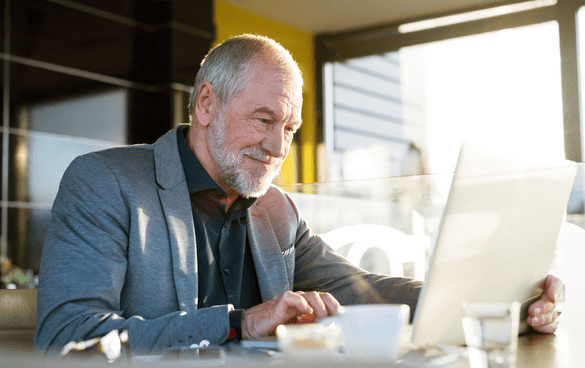 Older gentleman in a gray suit jacket and black button up sitting at a table and scrolling on a laptop