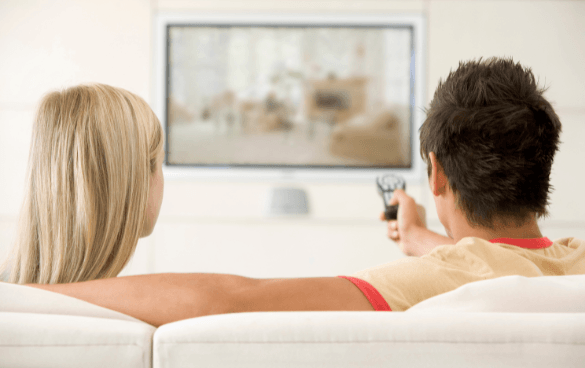 A man and a woman sitting on a couch from behind, watching a television screen, with the man holding a remote control.