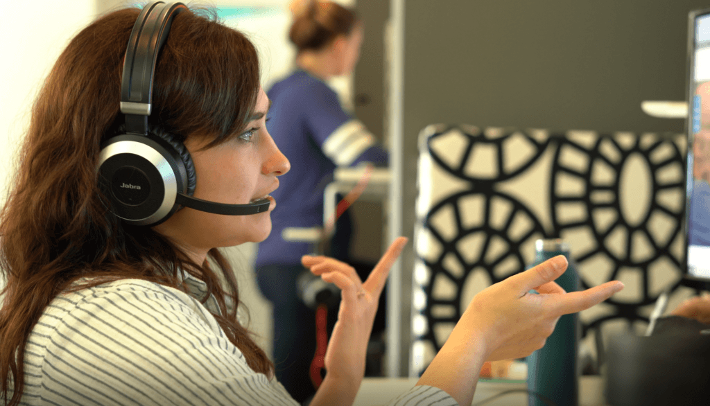 A woman in an office wearing a Jabra headset is speaking and gesturing with her hands, with a computer monitor in front of her and another person working in the background.